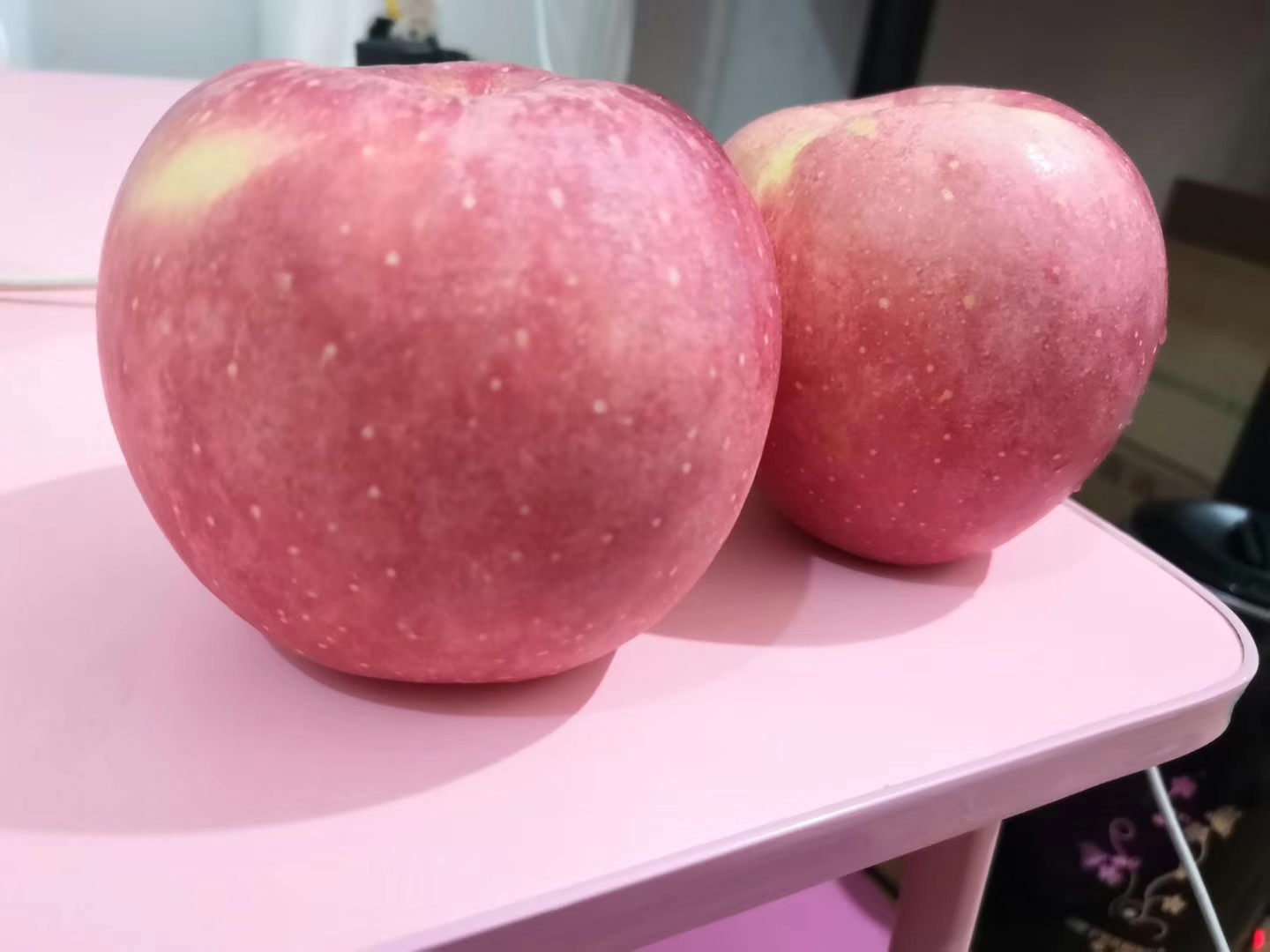 一个成熟的红富士苹果放置在木质桌面上,果皮呈现鲜艳的红色带有条纹光泽,表面光滑有自然蜡质层,背景为模糊的果园环境,阳光透过树叶洒在果实上 红富士苹果特写