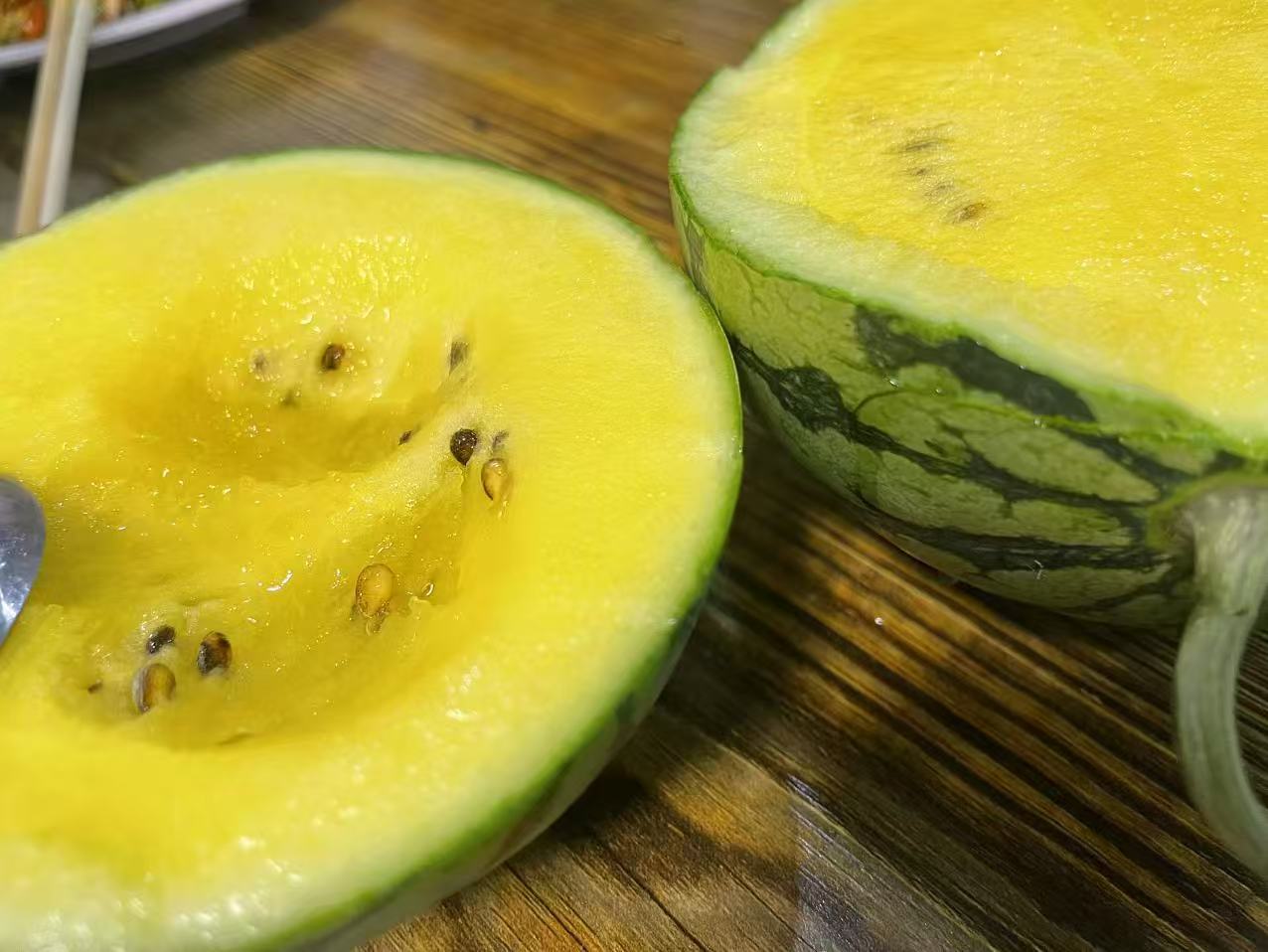 一个完整的特小凤西瓜被横向切开,露出金黄色的果肉和少量黑色籽粒,果肉水分饱满,表面有自然光泽,背景为田园风格的木制餐桌,周围散落着几片绿叶和水珠,展现清新自然的夏日氛围 特小凤西瓜切开图
