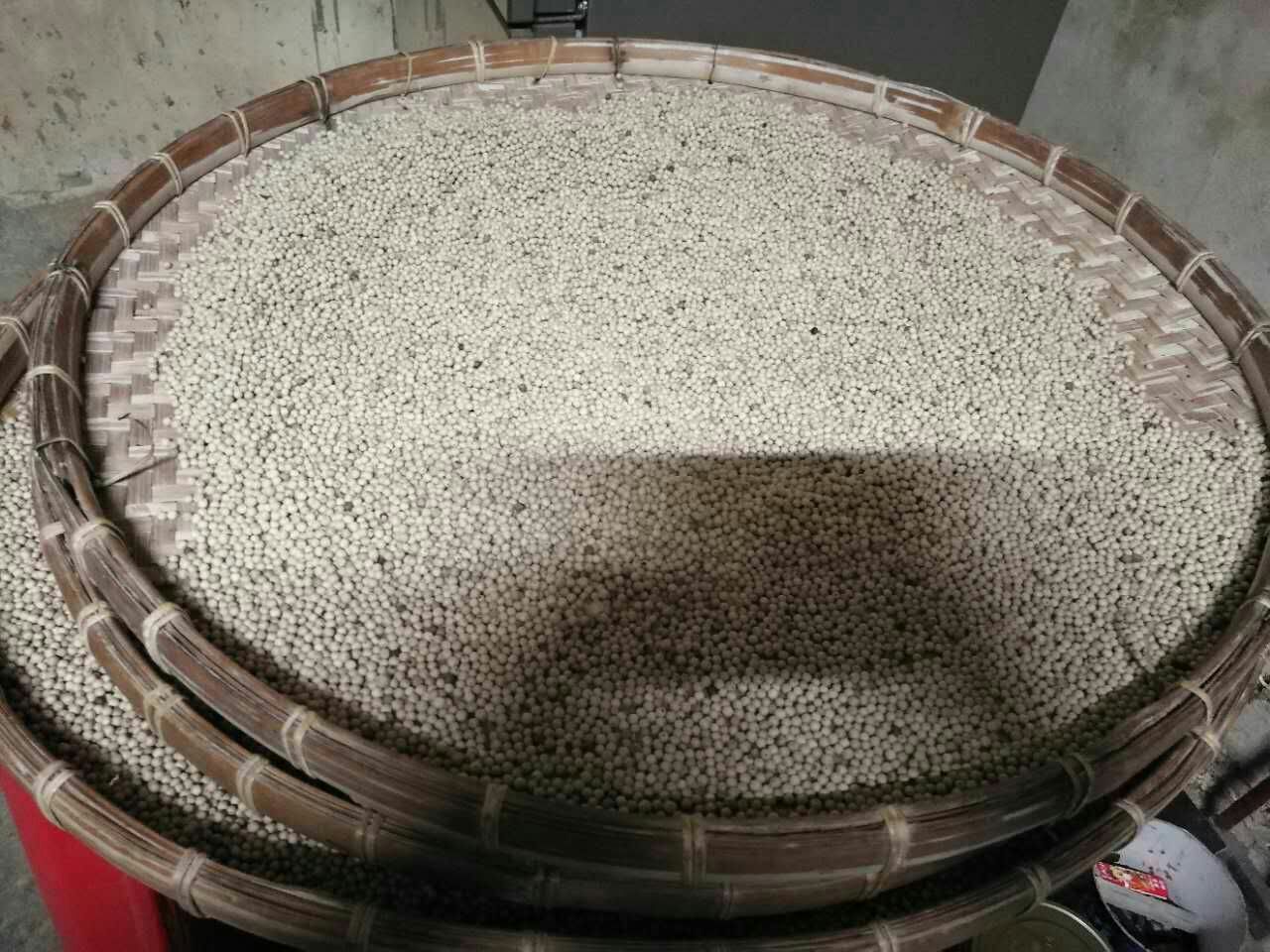一名身穿白色工作服的女工坐在竹编簸箕前,双手轻柔地翻动大量乳白色的白胡椒颗粒,背景是明亮整洁的加工厂内部,阳光透过窗户洒落在桌面上,凸显出白胡椒洁净的外观和精细的人工分拣过程 工人手工筛选白胡椒干粒