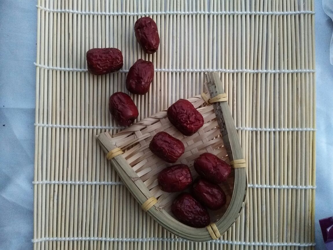 一位身穿蓝色布衣的农妇坐在竹编簸箕前,双手正在仔细分拣一堆新鲜红枣,将破损和颜色异常的果实挑出,背景是传统的农家院落,墙上挂着玉米和辣椒,阳光洒在簸箕上,营造出温馨而真实的乡村生活氛围 一位农妇正在手工挑选新鲜红枣