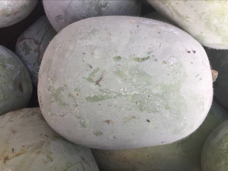 画面展示一个完整的白皮冬瓜放置在木质案板上,旁边有一把不锈钢刀正在削下白色的冬瓜皮,新鲜的冬瓜皮呈半透明状,表面带有细小绒毛,背景为自然光线下的厨房环境 新鲜白皮冬瓜及去皮过程特写