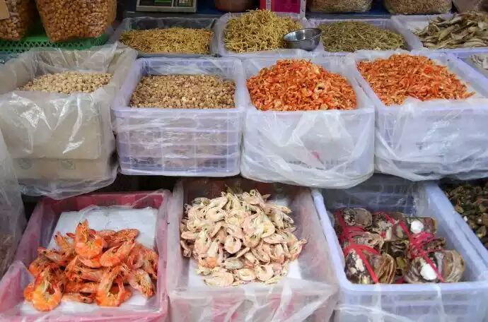 海鲜市场摊位实景,左侧冰台上陈列着整齐的新鲜对虾,虾体完整,色泽清亮;右侧保温箱内是真空包装的北极甜虾,标签注明‘加拿大进口’‘船冻熟制’。顾客正在咨询,摊主手持电子秤展示重量 市场选购实景图
