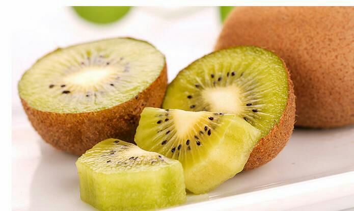 一个新鲜的海沃德猕猴桃被横向切开,露出翠绿色的果肉和排列整齐的黑色种子,果肉湿润多汁,周围有轻微水珠,背景为木质砧板,自然光线明亮柔和 猕猴桃切面特写