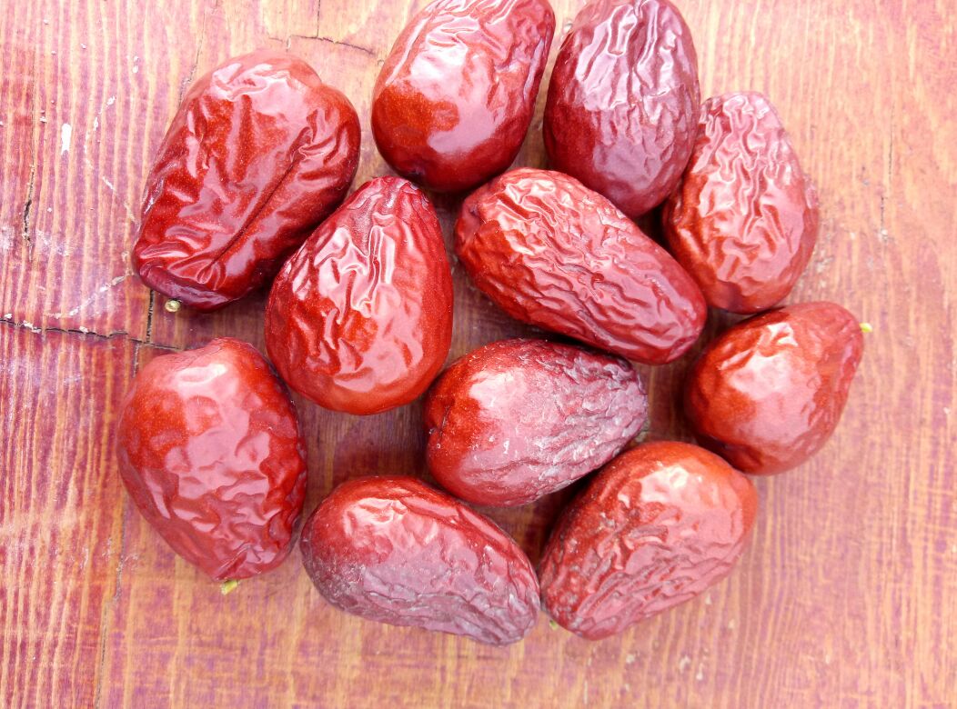 厨房场景中一名女性正在将骏枣切片加入正在熬煮的小米粥锅中,灶台上有蒸汽升起,旁边放着玻璃罐装的整颗骏枣和切片骏枣,背景为明亮的厨房环境,突出食材天然健康的氛围 骏枣应用场景图