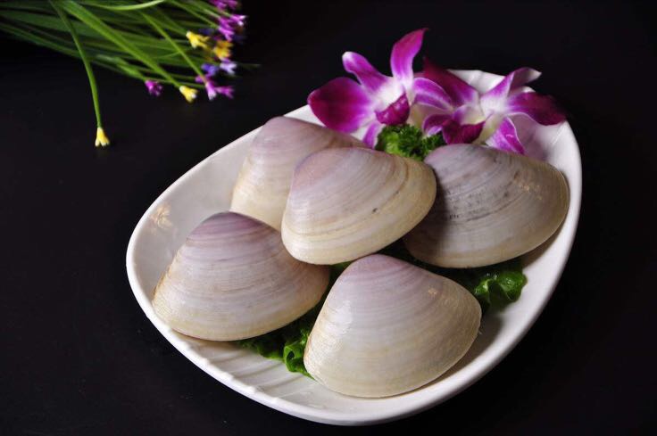 一位中年女性在海鲜摊位前弯腰挑选花蛤,摊主在一旁协助。台面上摆放着多个塑料筐,装满湿润的花蛤,背景可见其他海鲜品类。光线明亮,体现卫生整洁的市场环境 市场中消费者正在挑选花蛤的场景