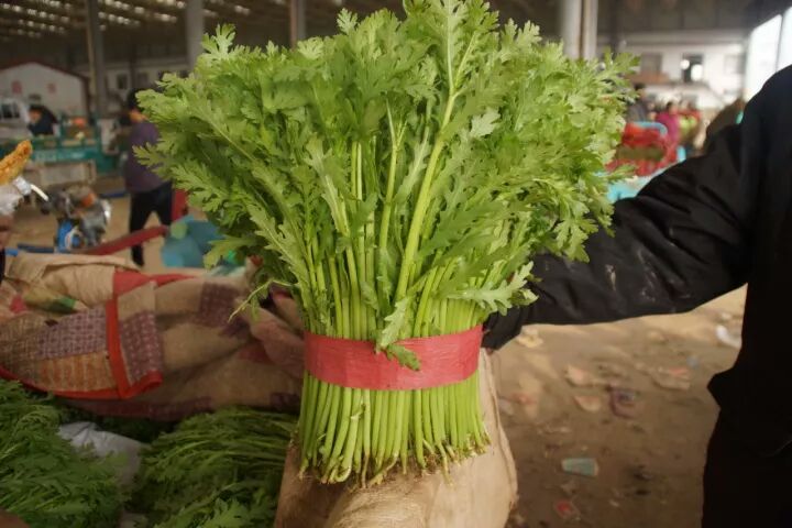 新鲜采摘的蒿子杆成捆堆放于菜市场摊位上，绿色茎秆粗壮挺直，叶片鲜绿，背景有秤和购物袋