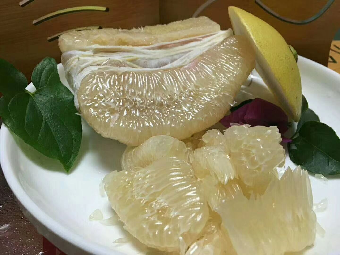 一个新鲜的红心柚被横向切开,露出粉红色的果肉和饱满的汁囊,外皮厚实呈淡黄色,果肉晶莹剔透,周围有几片剥好的柚子瓣放在白色瓷盘中,背景为自然木质餐桌,光线柔和,突出食材的新鲜与营养感 柚子切开特写