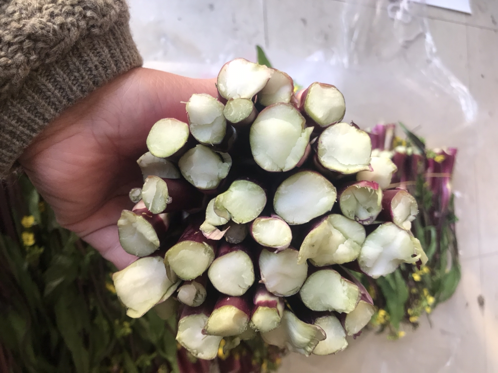 一位厨师正在厨房操作台上对新鲜红菜苔进行清洗,随后用刀将其斜切成段。部分已削去外层老皮的菜苔露出内部嫩红色的芯,旁边摆放着削皮刀和砧板,背景为整洁的中式厨房环境 红菜苔清洗与切段处理过程