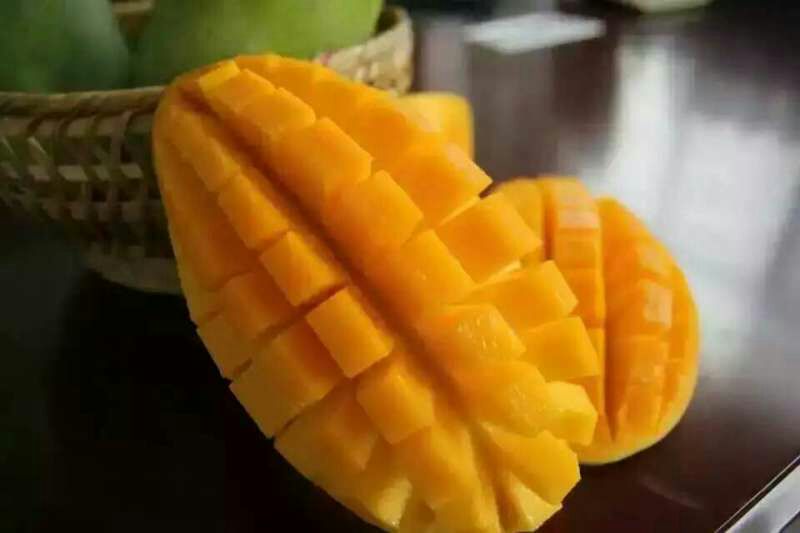 一位女性正在厨房中用刀将一颗青绿色的大青芒对半切开，露出金黄色的果肉和中间薄薄的果核