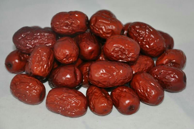 一位经验丰富的维吾尔族老匠人正坐在阳光充足的庭院中,手持弯月形牛角划枣刀,专注地在一颗饱满油亮的鲜骏枣表面进行纵向轻划;枣体呈深红近紫褐色,表皮光滑泛蜡质光泽,刀锋所至处渗出细微糖汁,背景可见竹匾、晾架及散落的干枣,整体光影温暖,凸显手工温度与地域特色 骏枣传统划枣工艺特写