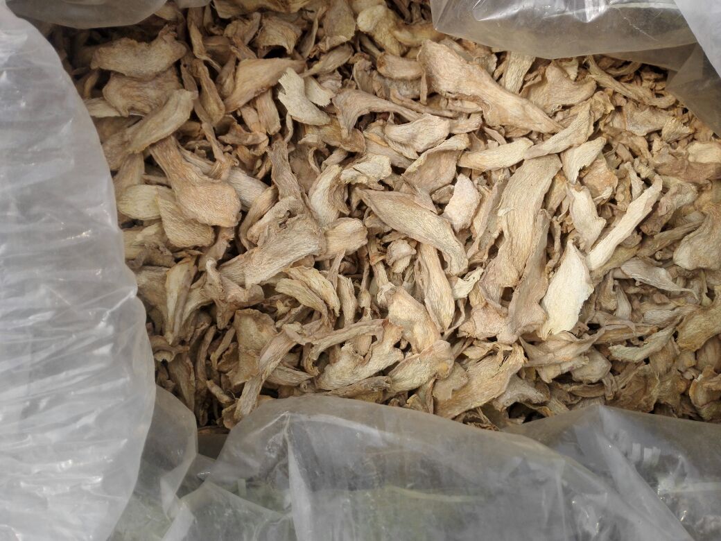 传统中药柜台内摆放着透明玻璃瓶,瓶中盛放干姜饮片,呈不规则薄片状,颜色棕褐,表面皱缩。标签上写着“干姜 50g”,背景可见其他中药材抽屉柜和戥子秤,体现干姜作为药材的专业使用场景 中药材铺中的干姜饮片展示