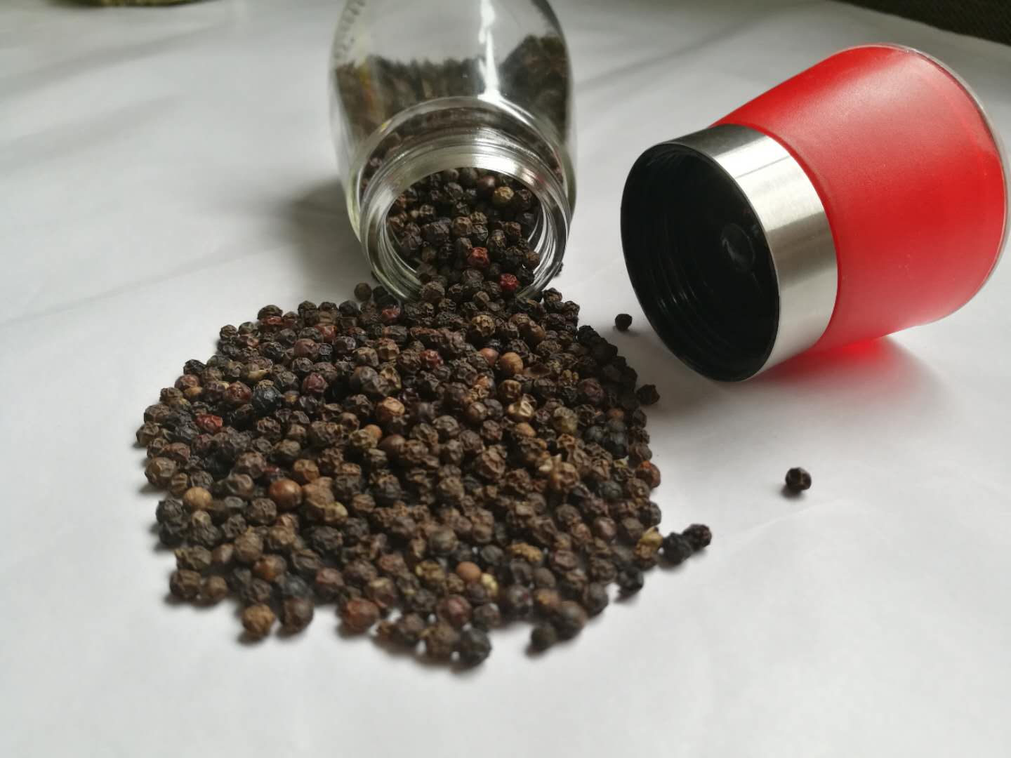 一张现代厨房场景照片:左侧为不锈钢胡椒研磨器,内装黑色颗粒;中间为玻璃瓶装红色辣椒粉;右侧为散落的整粒黑胡椒与碎辣椒段,背景为木质料理台面 厨房中常见的胡椒研磨器与辣椒粉瓶
