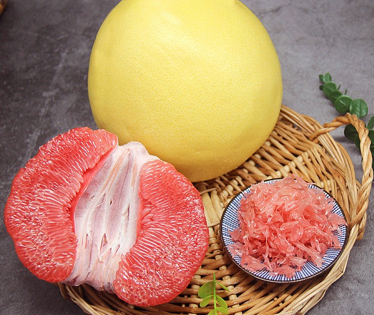 红心柚切开后露出鲜艳的红色果肉，果粒饱满多汁，周围摆放完整的红心柚果实，背景为果园环境