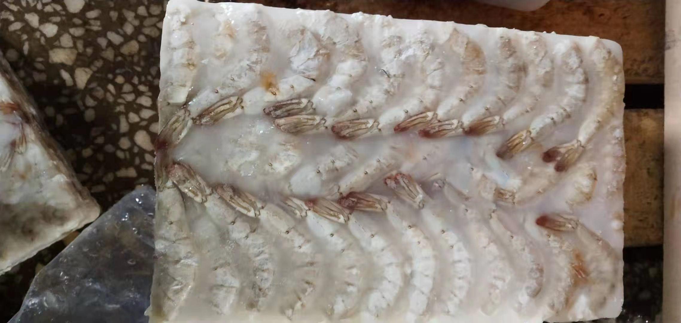 海鲜市场摊位上摆放着一盘冰鲜的红尾对虾,虾体整齐排列,覆盖着碎冰,背景有顾客正在挑选,摊主佩戴手套整理货物,环境整洁,体现真实消费场景 市场中红尾对虾售卖场景