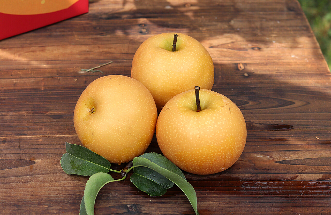 一张木质餐桌上摆放着三种水果:左侧为库尔勒香梨,中间为苹果,右侧为橙子,香梨旁放置一张标签写着‘库尔勒香梨|清热润燥’,背景虚化,突出香梨在清热类水果中的代表性地位 香梨与其他水果对比摆放