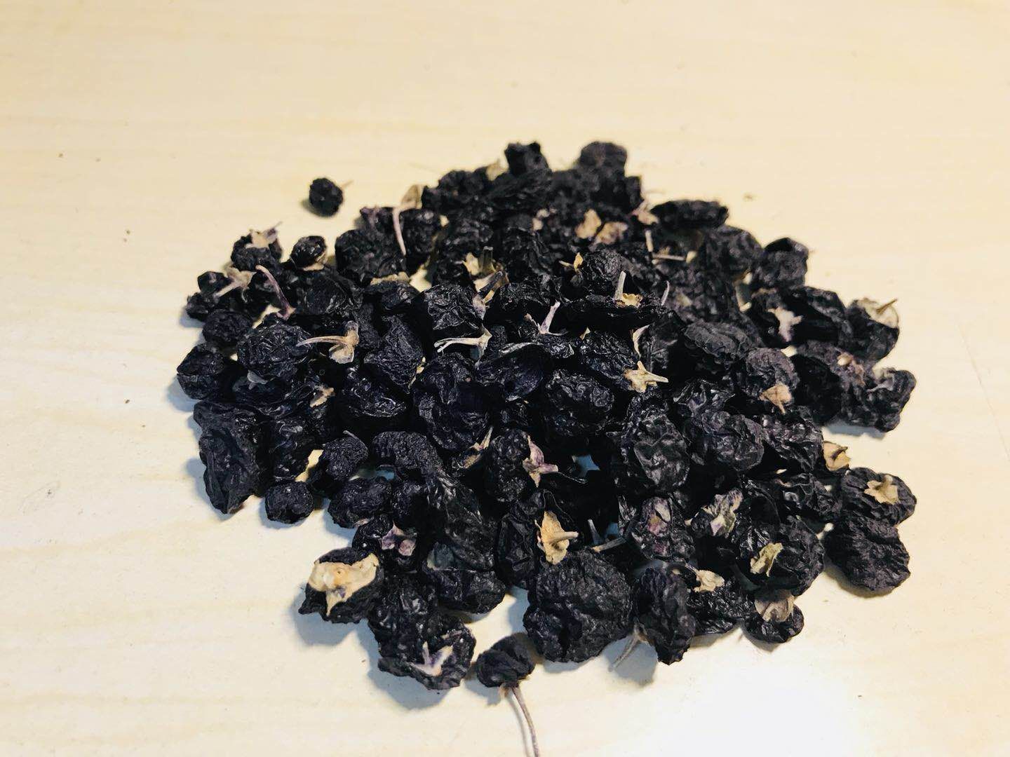 一簇干燥的黑枸杞果实,呈深紫黑色椭圆形颗粒状,表面有细微皱褶,背景为浅色木质桌面,旁边放有一把不锈钢茶匙,用于展示尺寸比例 黑枸杞特写镜头