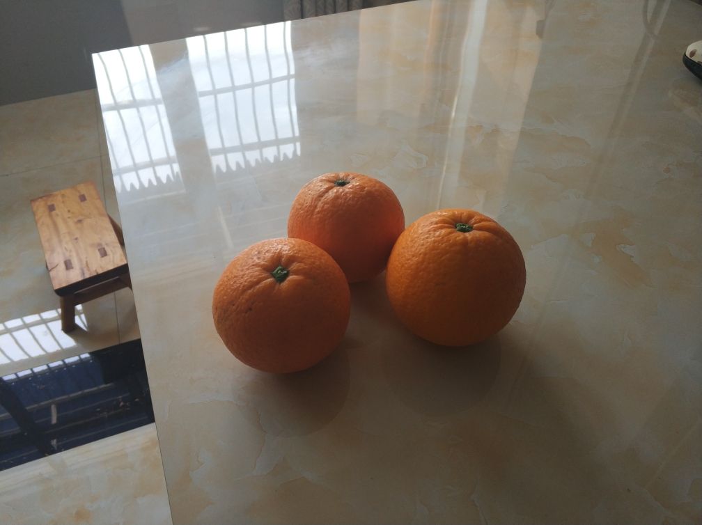 温馨家居摄影:原木餐桌中央铺着靛蓝扎染桌布,摆放三枚不同成熟阶段的秭归脐橙——左为初着色浅橙果(表皮微泛红晕),中为全熟金橙果(油胞细腻、光泽温润),右为晚熟深橙果(顶部略带玫瑰红斑);旁边配有陶瓷果盘、不锈钢柑橘勺、玻璃水杯中盛有澄澈橙汁,背景虚化处可见窗台绿植与‘屈原故里·脐橙之乡’木质挂牌 秭归脐橙家庭品尝场景