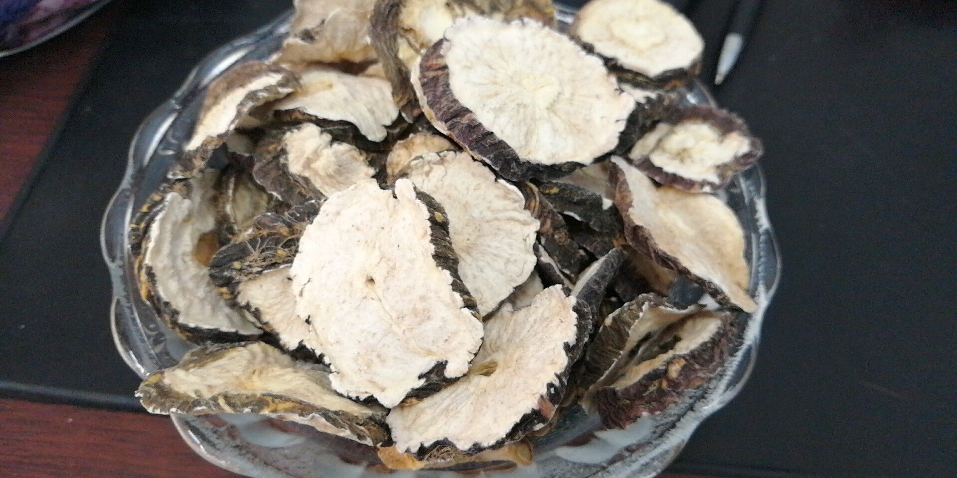 画面左侧为干燥的黑色玛卡切片,表面粗糙有纹理;右侧为经温水浸泡4小时后的玛卡片,体积略有膨胀,颜色变深,质地变软,边缘略透明。背景为木质砧板,旁边放置一个玻璃杯,杯中有淡黄色液体 黑玛卡干片与泡发后的对比图