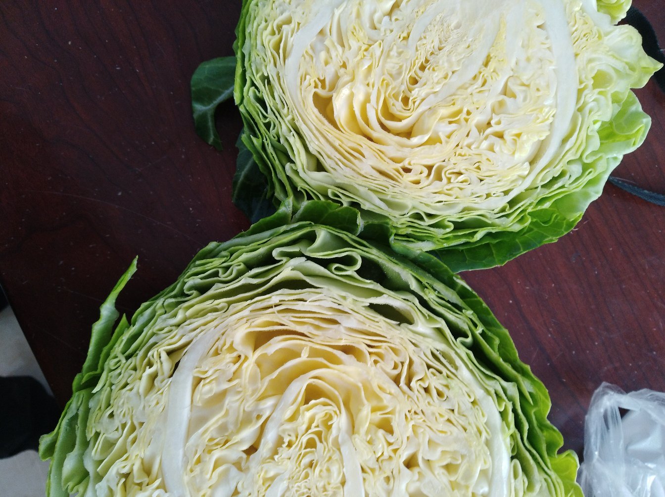 一颗新鲜的青甘蓝被横向切开,露出层层包裹的翠绿色叶片,切面紧实有光泽,背景为木质厨房台面,旁边有一把不锈钢刀 青绿色紧实的甘蓝切面特写