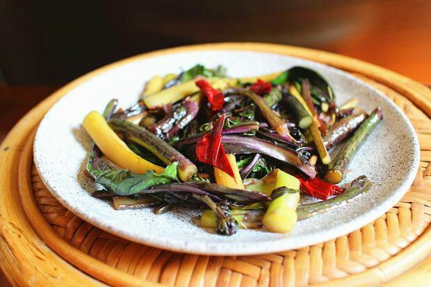 红菜苔家常烹饪场景：青花瓷盘中盛放清炒红菜苔，茎段翠紫相间、油亮诱人，配蒜末与红椒丝，背景为中式厨房台面，可见铁锅与木质砧板，整体色调温暖清新