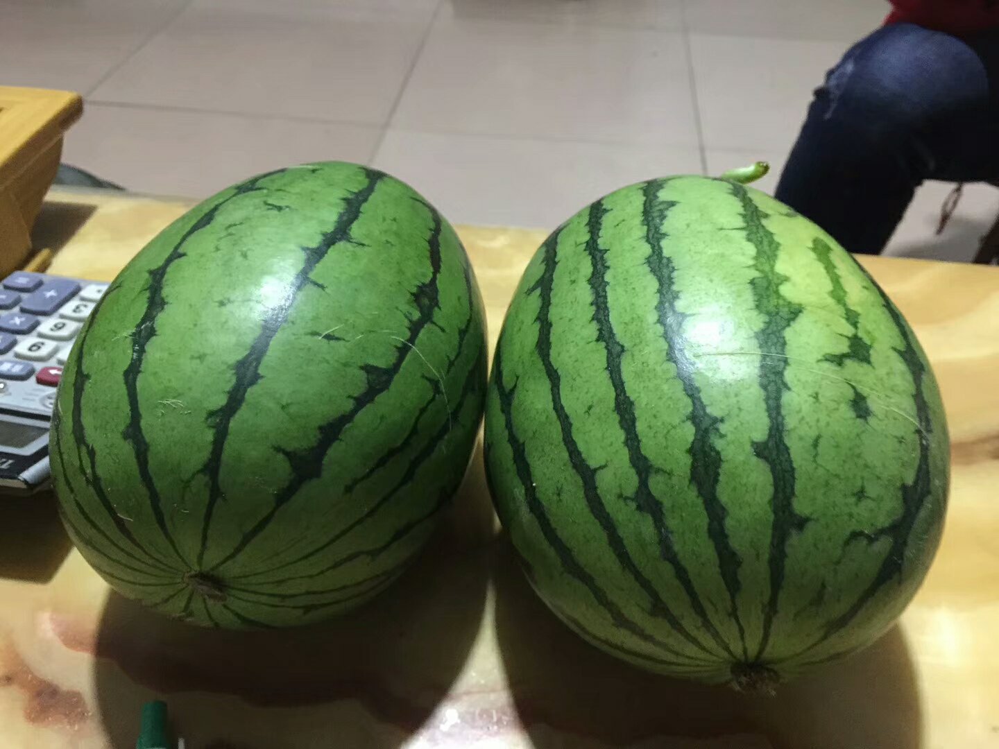 一个完整的红小玉西瓜放置在木质桌面上,瓜形圆润,表皮为翠绿色带有均匀的深绿色纵向条纹,背景虚化的厨房环境显示自然光线明亮,旁边有一把银色水果刀作为尺寸参照物 红小玉西瓜整体外观特写