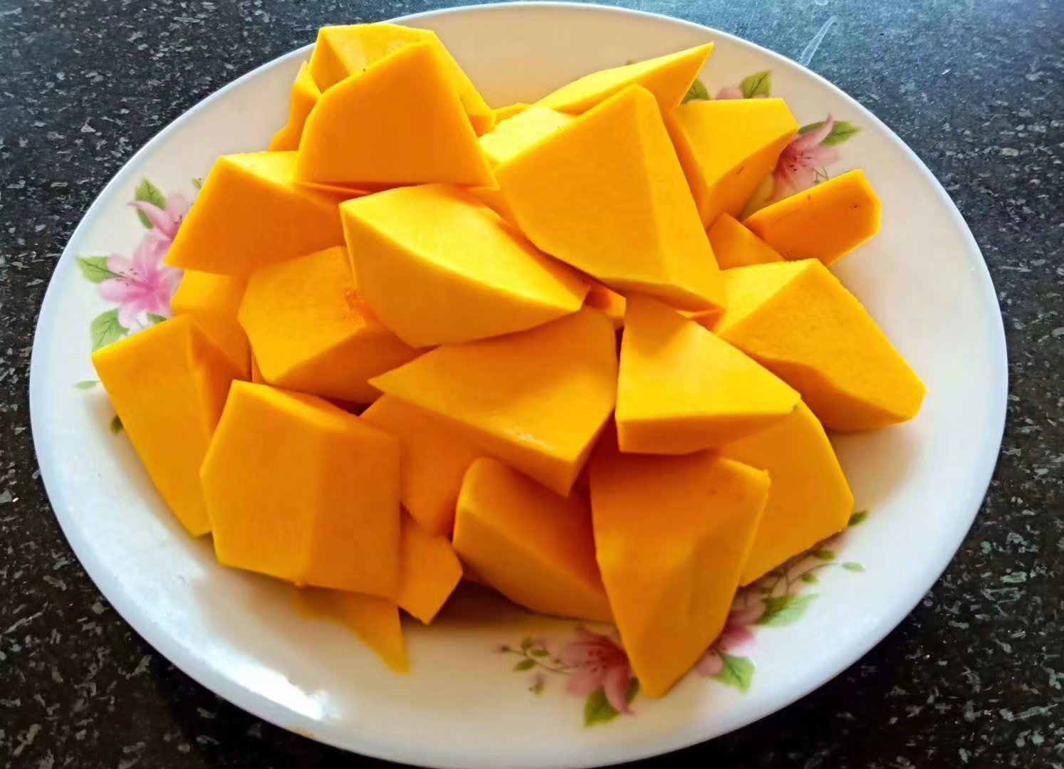 厨房操作台上摆放着切好的新鲜南瓜块、奶油瓶、搅拌锅和木勺，背景为开放式厨房环境，光线明亮温馨