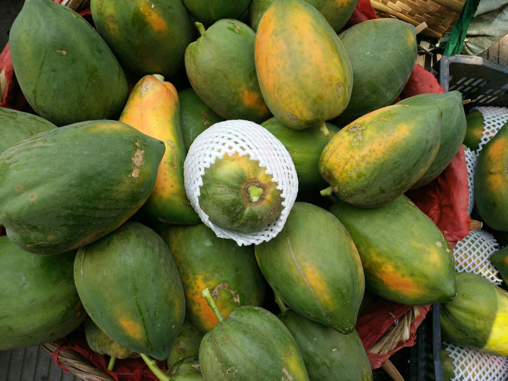 农贸市场摊位上整齐摆放着不同大小的成熟番木瓜，背景有消费者正在挑选，阳光透过棚顶洒落在鲜亮的果皮上