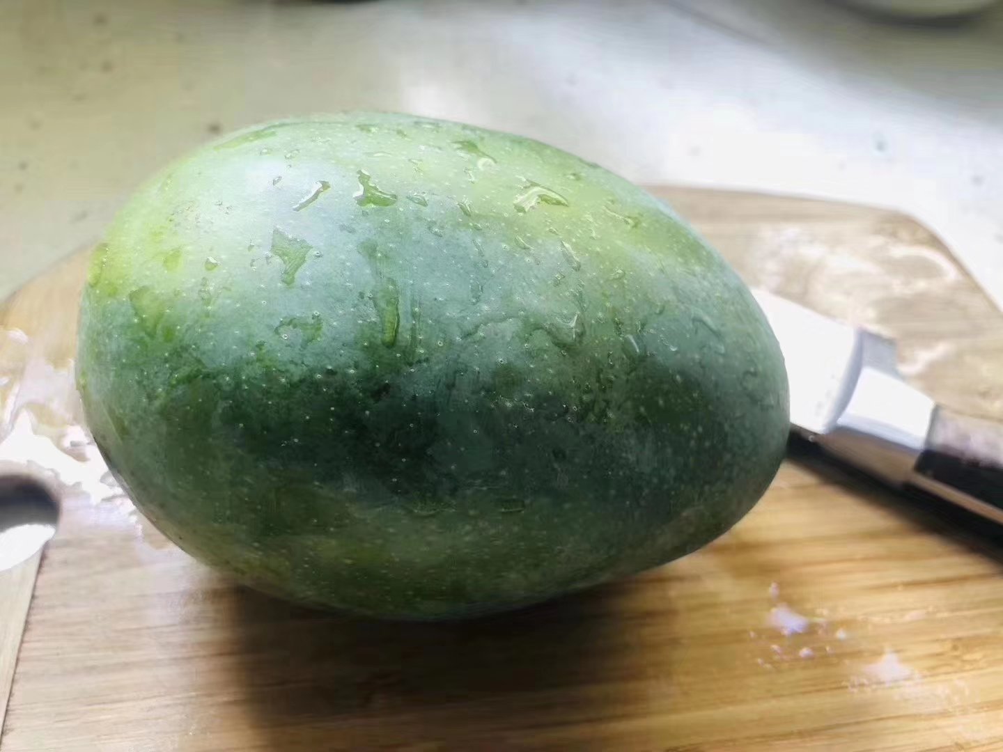 左侧为凯特芒,呈卵圆形,果皮深绿色;右侧为鹰嘴芒,尾部尖突,果皮青中带黄,整体较小巧。两者并列摆放于木质砧板上,背景为自然光下的厨房环境 凯特芒与鹰嘴芒外观对比图
