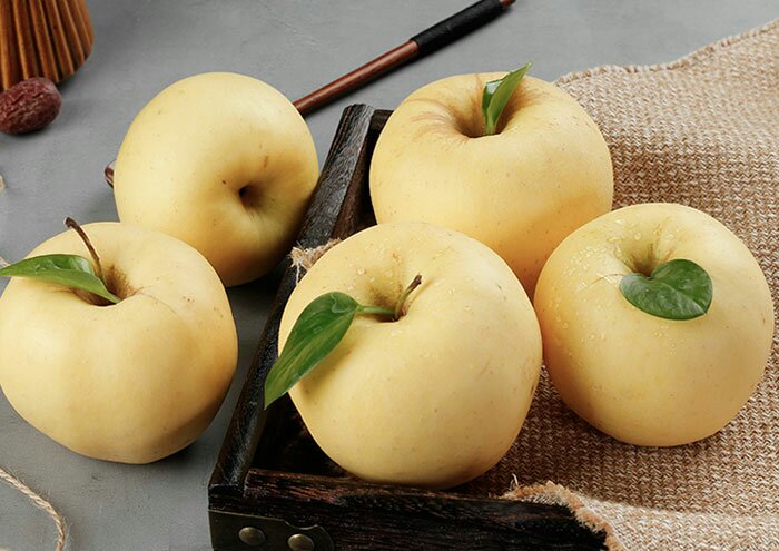 一篮新鲜采摘的黄元帅苹果摆放在木制桌面上，背景为果园风光