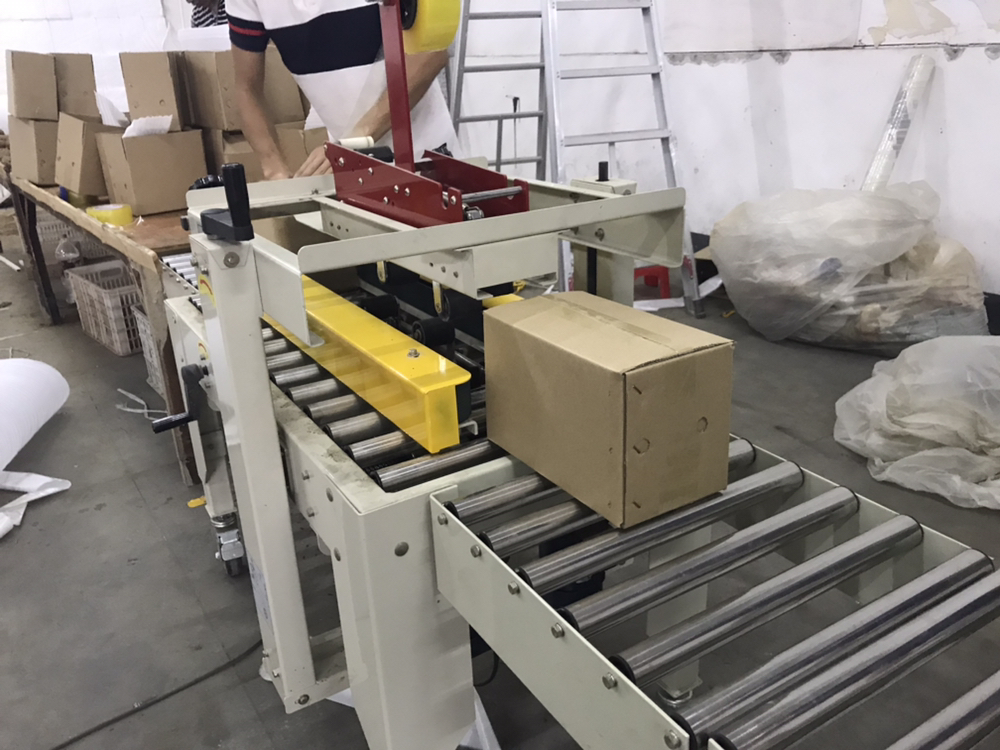一名工作人员正在仓库中将紫薯放入带有缓冲气泡膜的纸箱内,旁边堆放着印有品牌LOGO的包装箱,背景可见货架上的其他农产品包裹,展示规范化的电商发货流程,强调商品保护与专业包装 电商打包发货场景中的紫薯包装过程