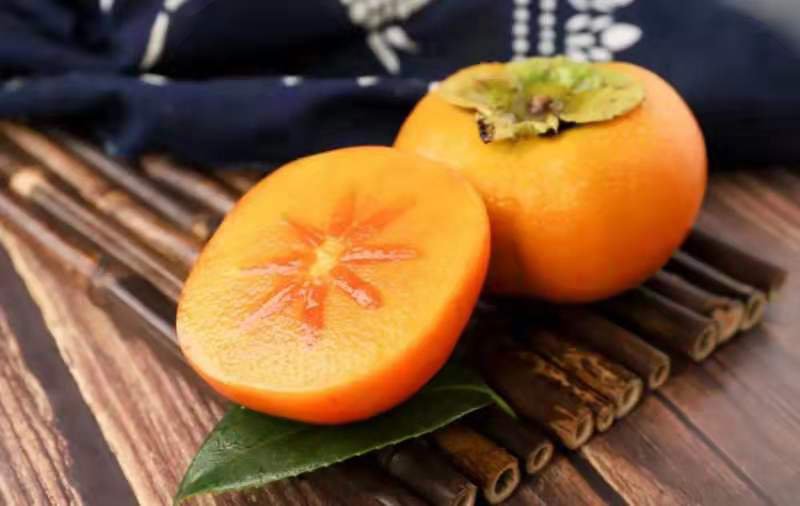 一颗成熟的巧克力甜柿,表皮呈橙红色,光滑有光泽;切开后果肉为深褐色,类似巧克力颜色,果肉细腻无籽,背景为自然果园环境,阳光柔和 巧克力甜柿整果与切面特写