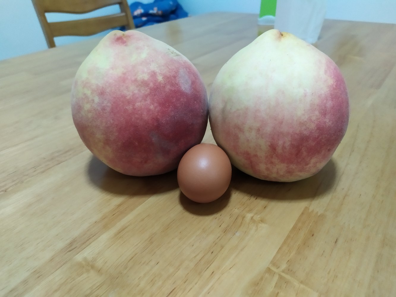 一个硕大的中华寿桃与两个普通水蜜桃并排摆放于木质托盘上,背景为果园环境,突出中华寿桃显著更大的体积和红润光泽的果皮 中华寿桃与普通桃对比图