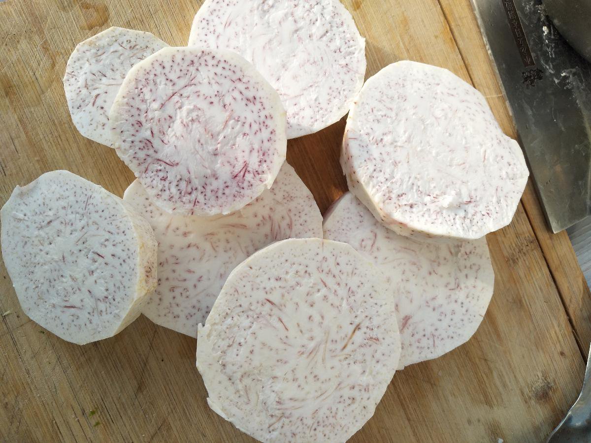 一位厨师佩戴手套正在案板上对一个紫红色外皮、肉质洁白的荔浦芋头进行去皮操作,旁边放着一碗清水,已切好的芋头块正浸泡其中,背景为厨房操作台 荔浦芋头去皮切块过程