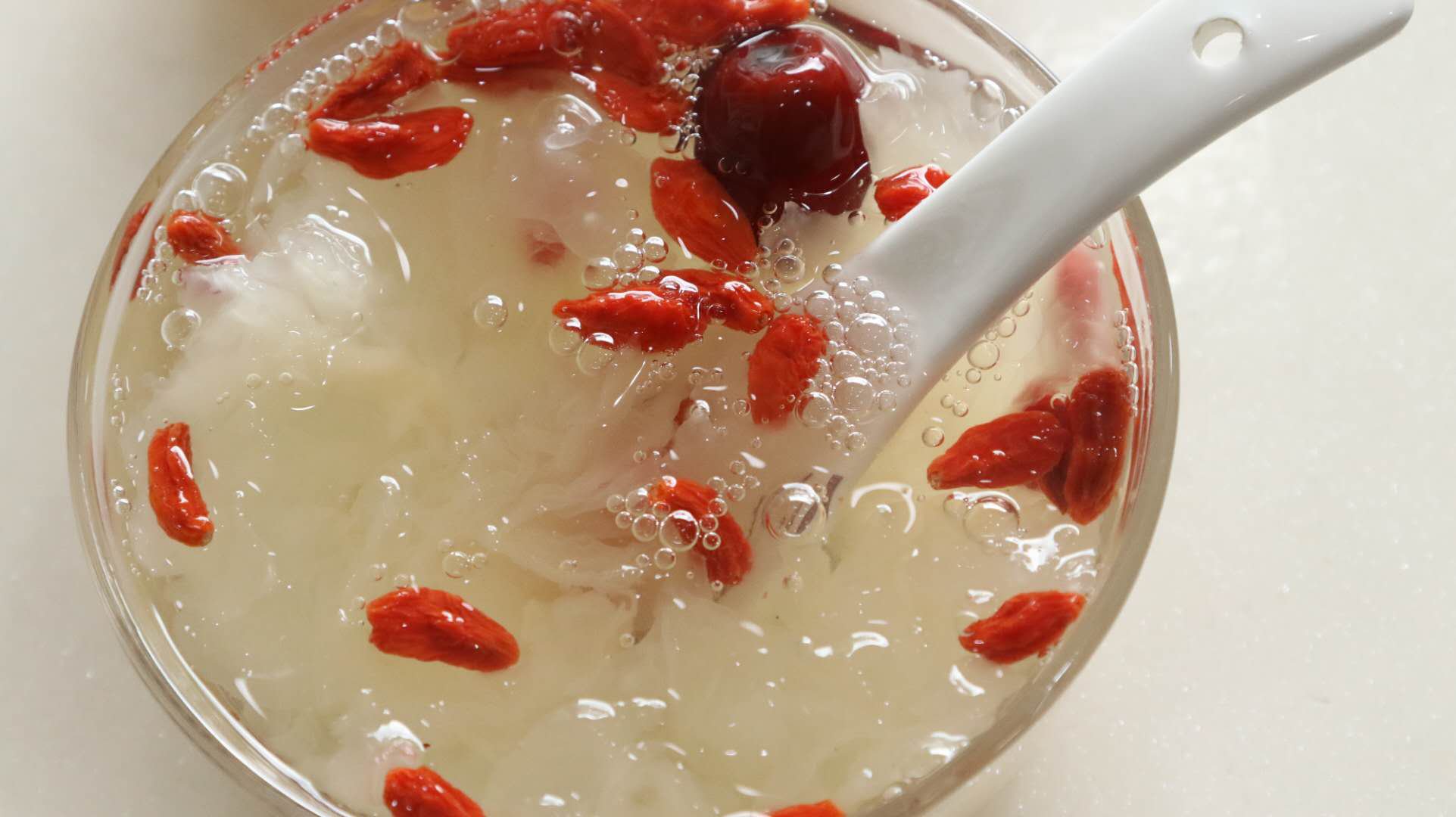 一碗刚出锅的椴木银耳羹,呈琥珀色半透明浓稠状,表面泛着油润光泽,勺子提起时可见拉丝现象,碗边附着细腻胶质,旁边点缀几颗枸杞和红枣,整体呈现温暖诱人的视觉效果 慢炖出胶的椴木银耳甜品特写