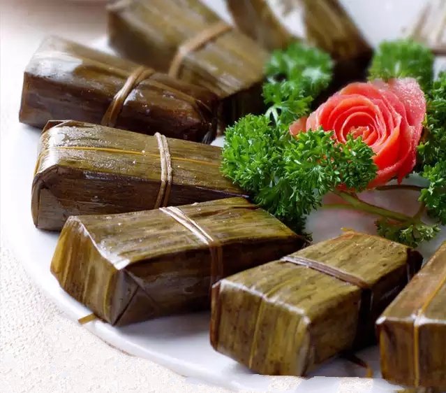 俯拍高清图:一只解开棉线的四角粽,粽叶翠绿微泛油光,剥开可见琥珀色糯米包裹着金黄咸蛋黄、棕红五花肉丁与碧绿蚕豆仁,豆粒完整分明,米粒晶莹油润,表面淋少许琥珀色酱汁,背景为原木砧板与青竹筷 刚出锅的青蚕豆五花肉粽特写