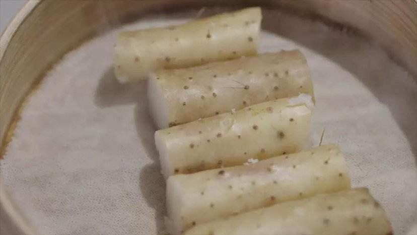 一位厨师正在厨房操作台上处理白玉山药,左手持山药段,右手使用削皮刀去皮,案板旁放有生姜片、红枣和枸杞,炉灶上的砂锅正冒着热气,显示正在进行炖煮 厨房中正在烹饪白玉山药的场景