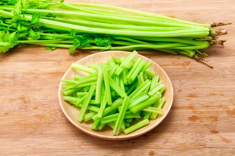 新鲜香芹整齐摆放在案板上,左侧为带老叶和粗筋的原始状态,右侧为已去除老叶、撕去纤维并斜切成段的处理后状态,旁边配有削皮刀、砧板和切好的蒜片 香芹处理前后对比图