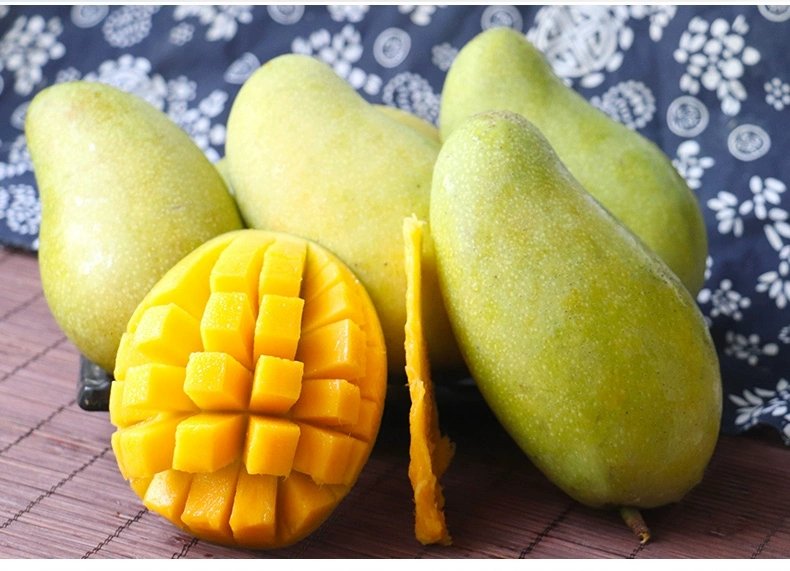 一颗完整的青绿色大青芒放置在木质桌面上,旁边是一颗被横向切开的大青芒,露出金黄色的果肉和光滑的果核,背景有轻微虚化的绿叶,展示其从外皮到果肉的颜色对比 大青芒整颗与切开对比图