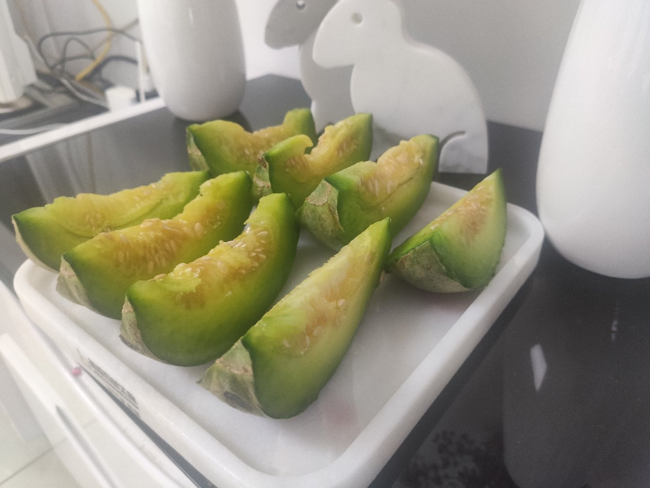 一份由绿宝甜瓜块、蓝莓、黄瓜片和薄荷叶组成的健康水果拼盘,摆放在白色陶瓷盘中,背景为厨房台面,旁边有一杯透明玻璃水杯,整体风格清新自然 绿宝甜瓜水果拼盘