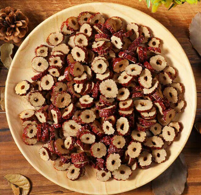 一盘摆放整齐的干红枣片,呈深红色,表面有自然褶皱,部分为切片状,背景为木质餐桌,旁边配有白色陶瓷碗和绿色植物,展现自然健康的氛围 红枣干特写