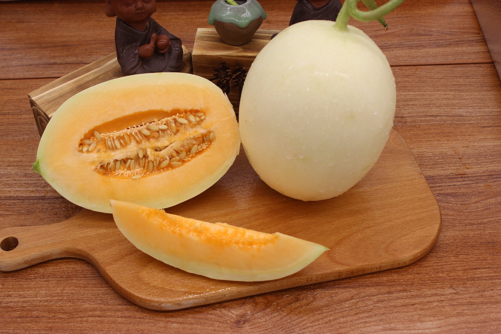 一颗完整的翠梨甜瓜放置在木质砧板上,果皮呈均匀青绿色,表面光滑无网纹,旁边切开一半展示内部洁白脆嫩的果肉和少量籽腔,背景虚化为阳光照射下的瓜田 翠梨甜瓜特写照片