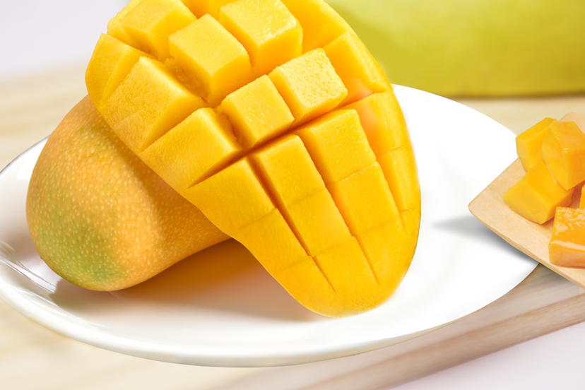 新鲜芒果切配场景：厨师手持锋利果刀，正将一枚金煌芒沿核对称剖开，果肉金黄油亮，可见数条纤细深褐色维管束自然延伸，砧板洁净，背景为明亮厨房环境