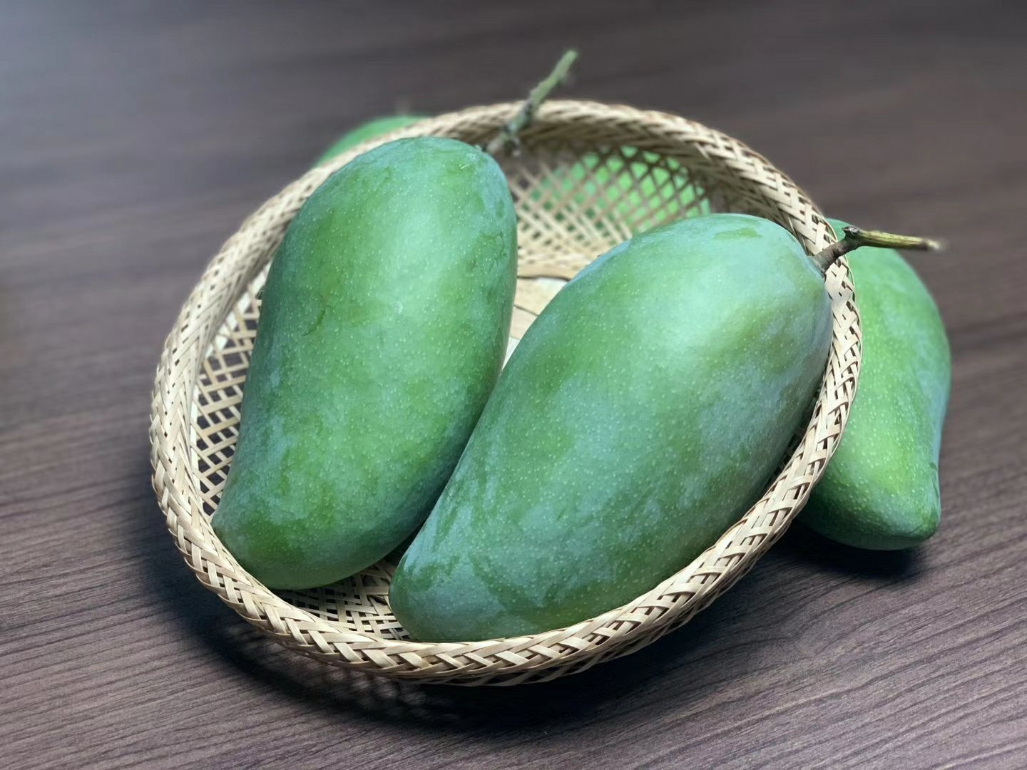 一个完整的青绿色大青芒放置在木质桌面上,表皮光滑呈椭圆形,背景为自然光线下的厨房环境,旁边有一把水果刀用于对比大小 大青芒整体外观特写