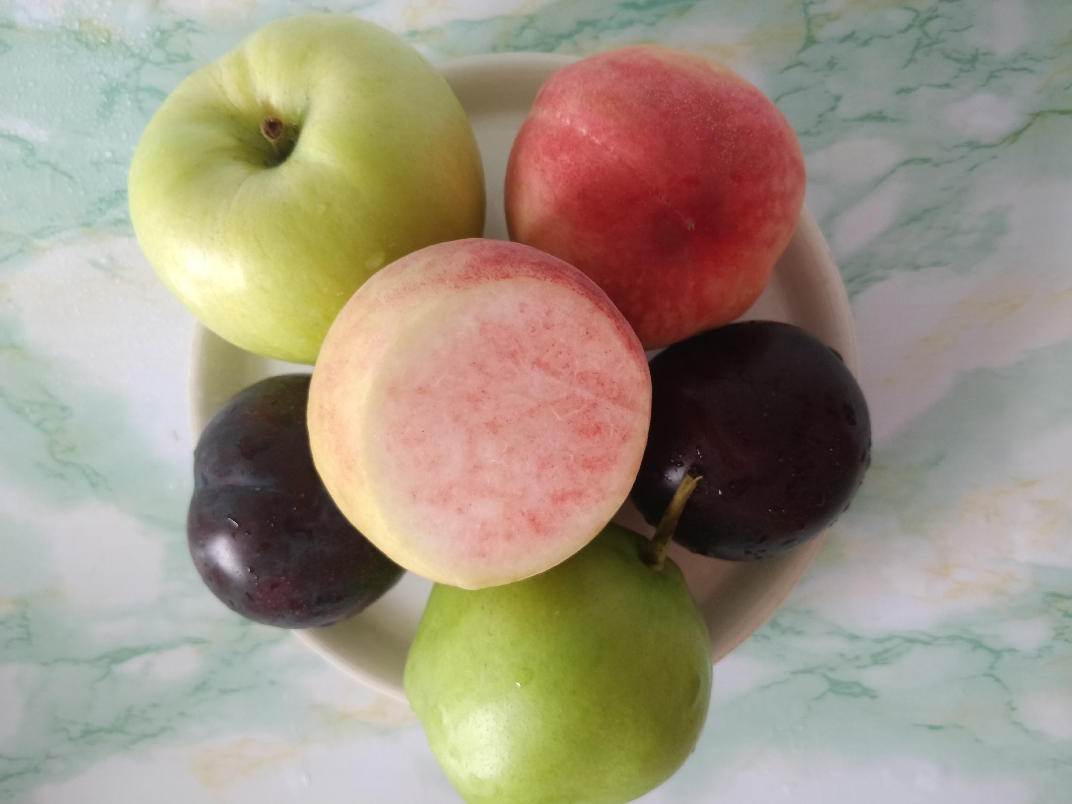 一张木质桌面上摆放着三种不同状态的黑布林李子:左侧是刚采摘的新鲜果实整齐排列在竹篮中;中间是切片后铺在白色烘焙纸上的李子片,准备风干;右侧是透明玻璃罐中装满深褐色的李子干,背景为厨房环境,体现家庭自制场景 黑布林李子保存方法示意图