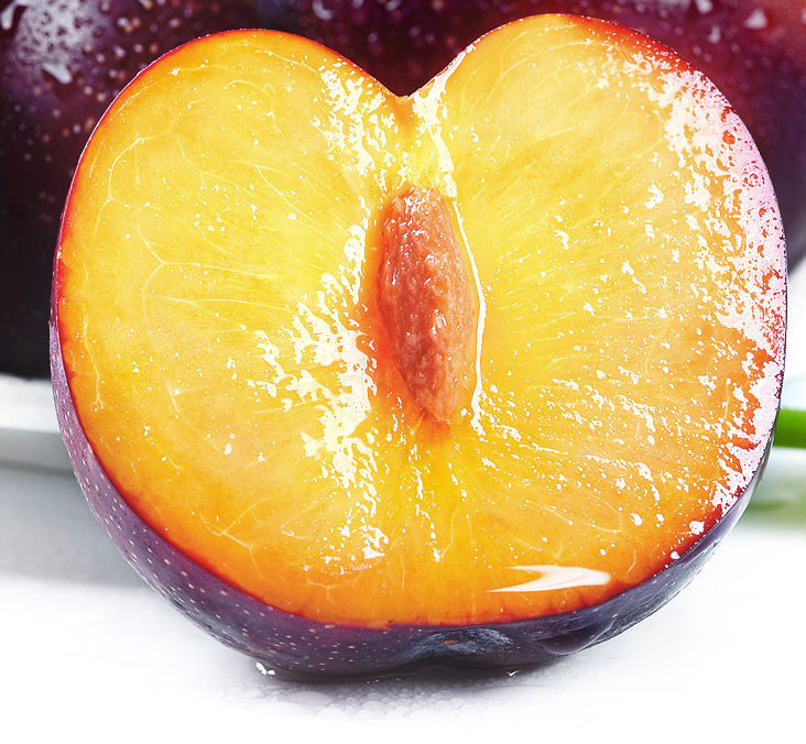 一个被横向切开的黑布林李子,露出金黄色至淡红色的果肉,果核清晰可见且较小,果肉纹理细腻,汁液丰富,旁边摆放着几颗完整未切的深紫色果实,背景为木质砧板和自然光环境,突出水果的新鲜与质感 切开的黑布林李子特写