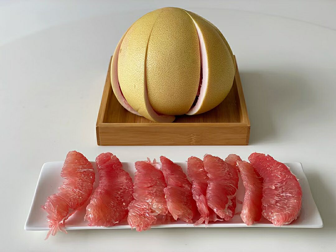 一个被横向切开的三红柚,露出鲜艳的玫瑰红色果肉,果粒分明、晶莹剔透,周围有几片剥好的果瓣放在白色瓷盘中,背景虚化的厨房台面上摆放着蜂蜜罐、柠檬片和玻璃杯,营造出健康生活的氛围 切开的三红柚特写