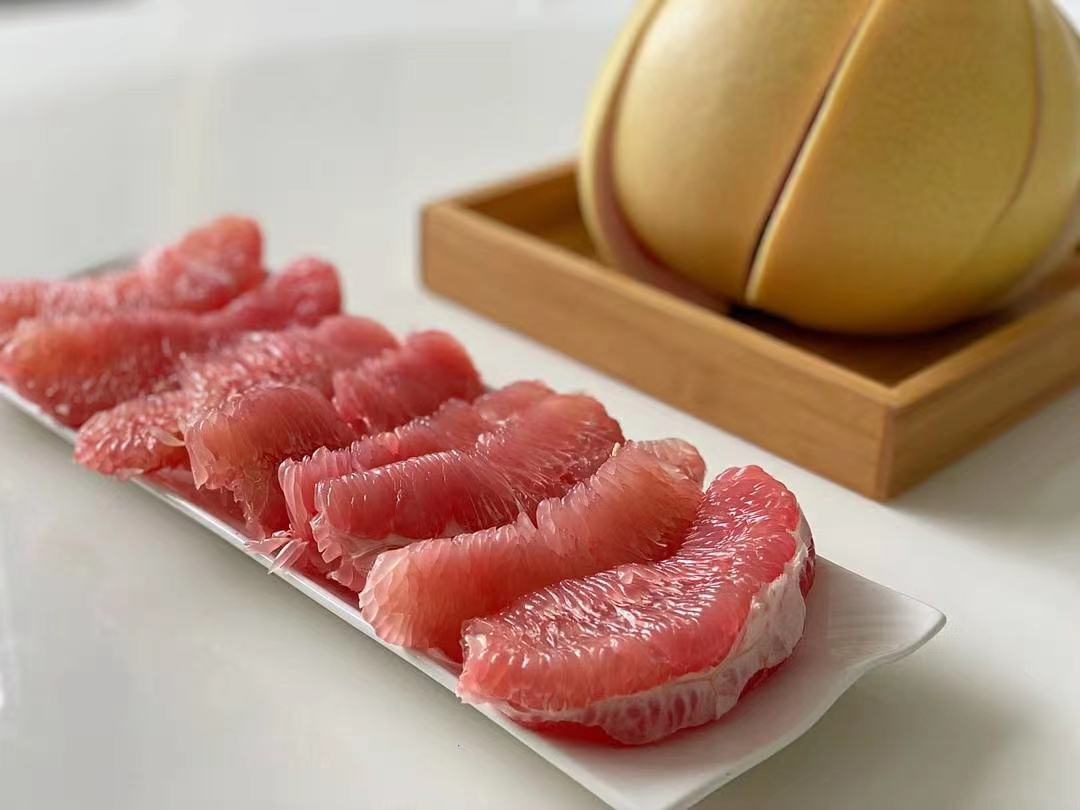 一位年轻女性坐在阳台的椅子上,手中拿着一瓣红心柚正在食用,背景是夜晚的城市灯光,桌上放着一杯温水和半个去皮的红心柚,营造出轻松健康的晚间生活氛围 女性夜晚在阳台吃柚子场景