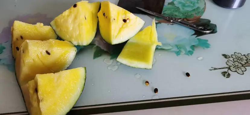 一个完整的特小凤西瓜被横向切开,露出鲜艳的金黄色果肉,果肉质地细腻均匀,中心区域有少量黑色成熟种子呈放射状排列,背景为浅色木质砧板,周围散落几片瓜瓣,光线柔和自然 特小凤西瓜切开图展示金黄色果肉与黑色种子分布