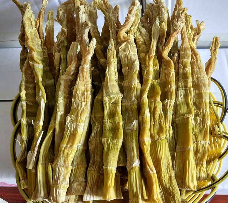 一堆刚挖出来的新鲜竹笋整齐地码放在竹编筐里,阳光洒在其上,旁边是一盘色泽金黄的传统手工笋干,展现从原料到成品的对比过程 新鲜竹笋堆放在竹筐中,旁边摆放着已制成的金黄色笋干,背景为乡村厨房场景
