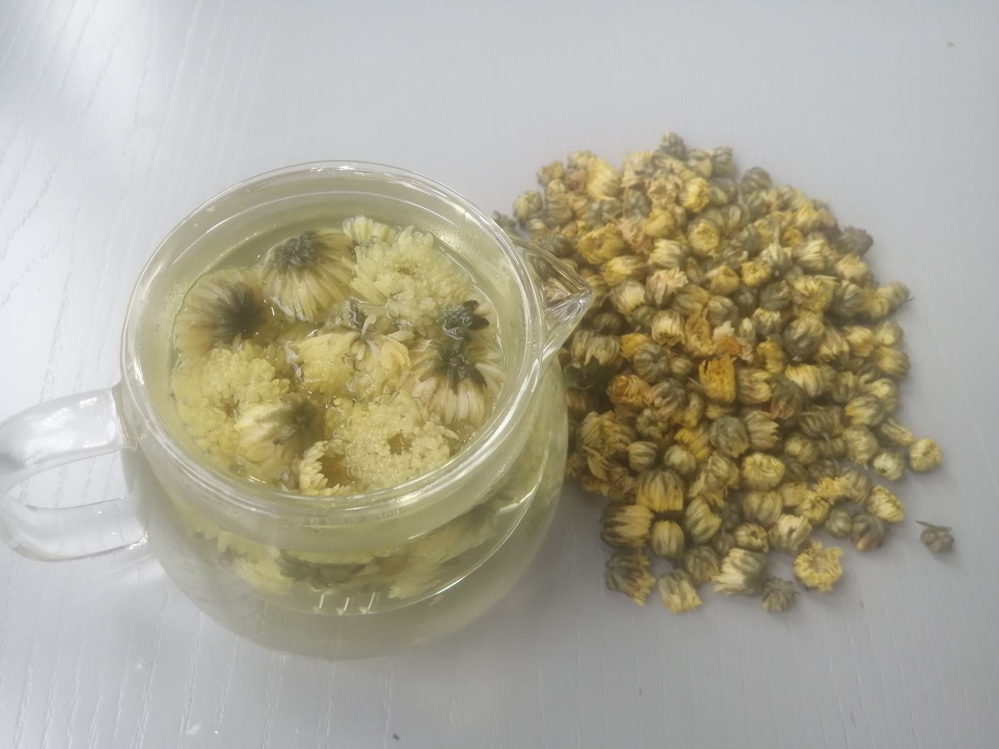 生活美学场景：素白陶瓷盖碗中盛放5朵胎菊王，注入90℃温水后微微浮沉，汤色澄澈透亮呈淡琥珀黄，几缕细微绒毛悬浮其中，旁置竹制茶则与手写“菊胎”书法签，整体构图宁静雅致